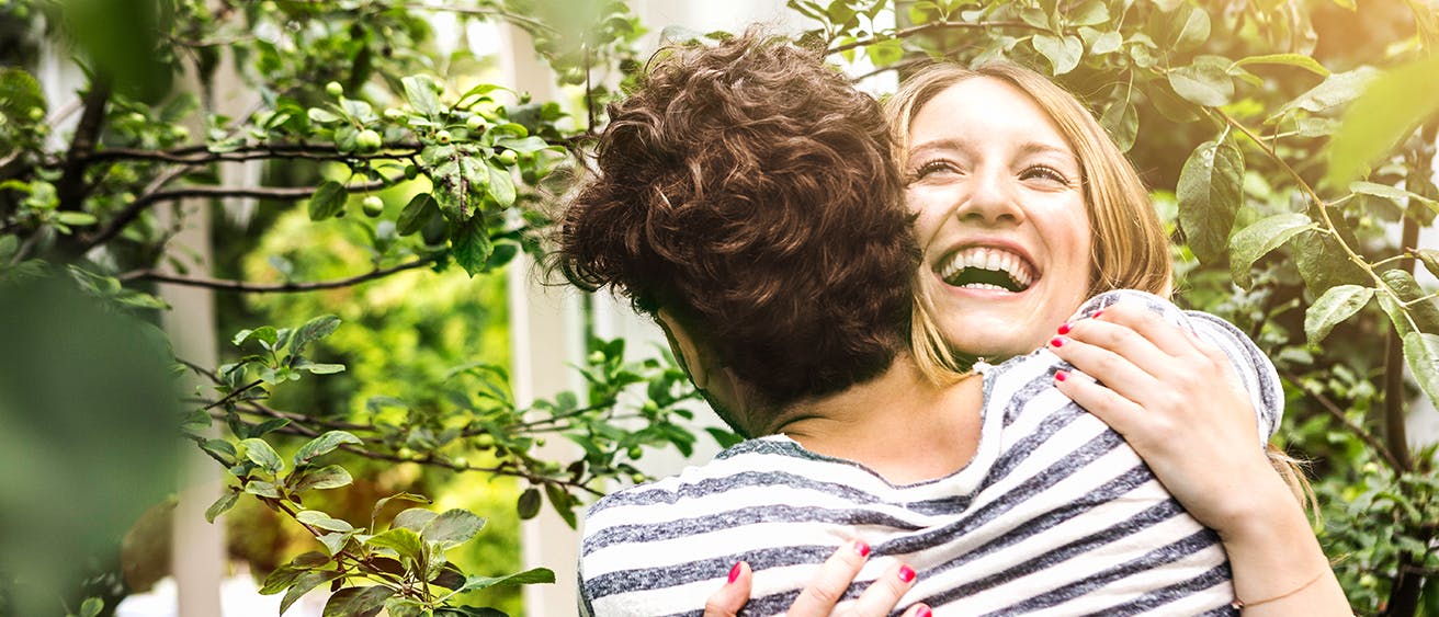 A smiling, young woman with blonde hair hugging a young dark-haired man surrounded by branches with leaves.